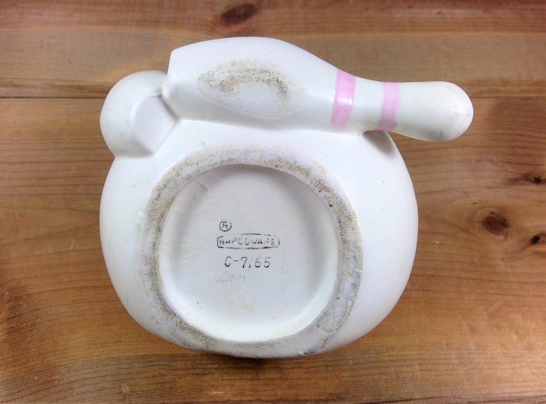 May include: White ceramic bowling pin shaped piggy bank with a pink stripe. The bottom of the bank has the text "(R) NAPCOWARE C-7765".