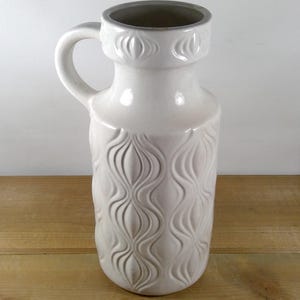May include: White ceramic vase with a textured, wavy design. The vase has a handle and a wide opening.