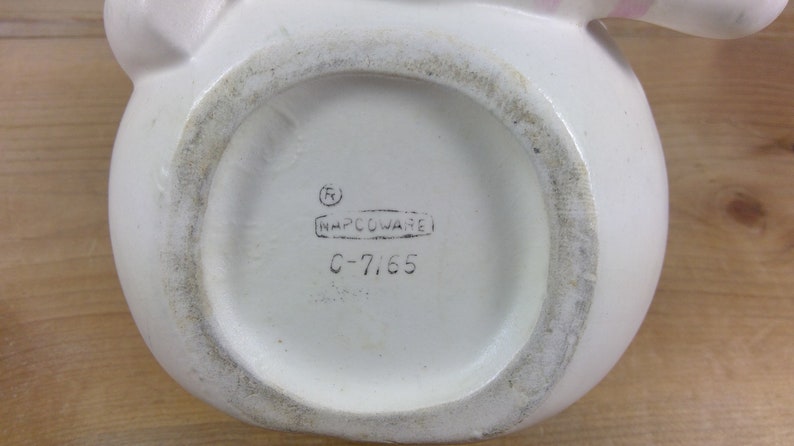 May include: White ceramic pitcher with a pink handle and a black stamp on the bottom that reads "(R) NAPCCOWARE C-7165".