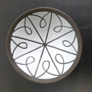 May include: A small, round, black ceramic dish with a white glaze. The dish features a decorative design of white lines forming a geometric pattern.