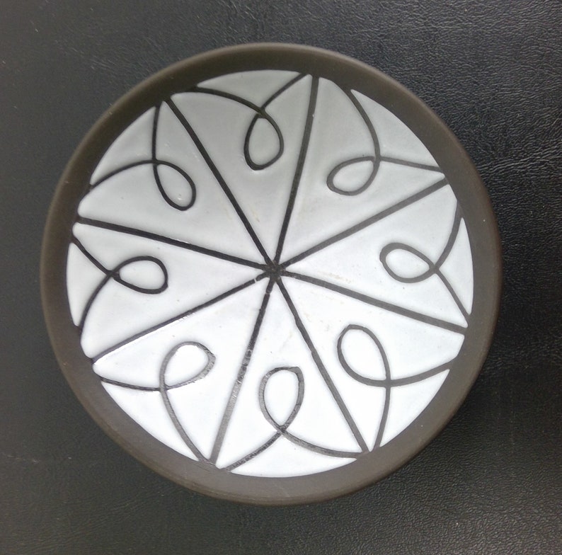 May include: A small, round, black ceramic dish with a white glaze. The dish features a decorative design of white lines forming a geometric pattern.
