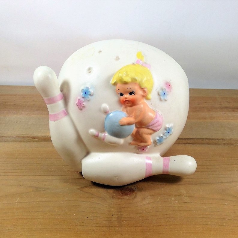 May include: A white ceramic wall planter shaped like a bowling pin with a baby holding a bowling ball. The planter is decorated with pink and blue stripes and flowers.