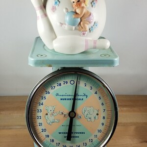May include: A blue and white baby scale with a white ceramic bowling pin with a baby girl figurine on top. The scale face reads "American Family Nursery Scale" and has a teddy bear and elephant illustration. The scale reads up to 29 pounds and 12 ounces.