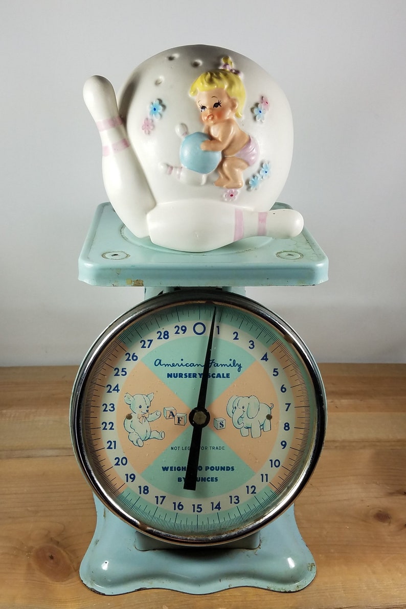 May include: A blue and white baby scale with a white ceramic bowling pin with a baby girl figurine on top. The scale face reads "American Family Nursery Scale" and has a teddy bear and elephant illustration. The scale reads up to 29 pounds and 12 ounces.