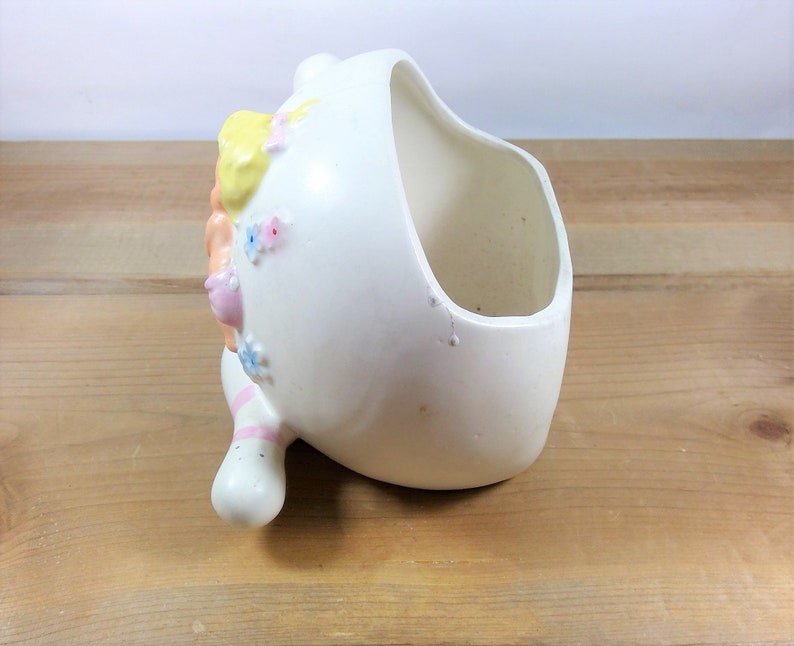 May include: White ceramic planter shaped like a girl's head with pink, yellow, and blue painted details. The planter has a wide opening for planting.