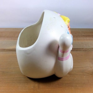 May include: White ceramic planter shaped like a bowling pin with a girl's face on the side. The planter has pink and blue stripes on the bowling pin.