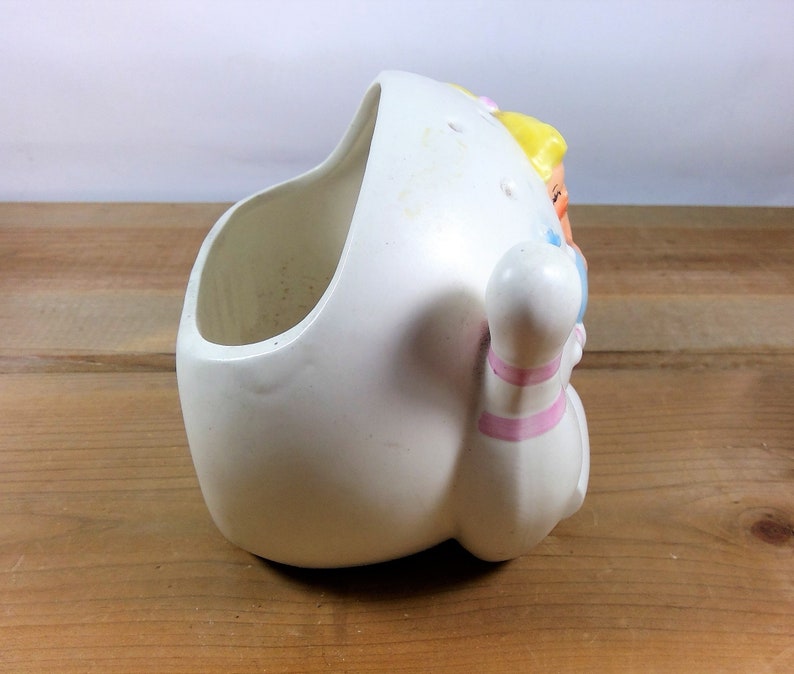 May include: White ceramic planter shaped like a bowling pin with a girl's face on the side. The planter has pink and blue stripes on the bowling pin.