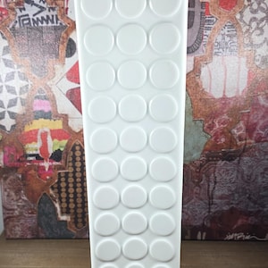 May include: A white ceramic vase, tall and rectangular, adorned with raised circular details. The vase has a smooth, glossy finish and is set against a colourful, abstract collage background. It sits on a wooden surface.