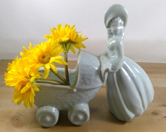 May include: A light blue ceramic baby carriage planter with a woman figurine holding the handle. The carriage has a textured surface and is filled with yellow flowers.
