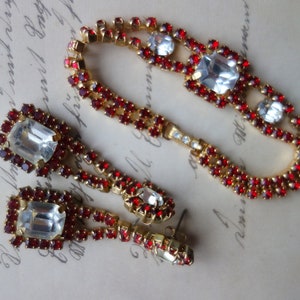 Könnte beinhalten: Ein Vintage-Schmuckset mit Armband und Ohrringen. Die goldfarbenen Stücke sind mit funkelnden roten und klaren Strasssteinen verziert. Die Ohrringe haben eine rechteckige Form, während das Armband ein geschwungenes Design aufweist.