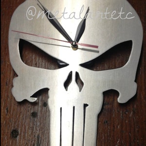May include: A silver metal wall clock shaped like the Punisher skull. The clock has black hands and a red second hand. The clock face is blank and the skull is cut out. The clock is mounted on a wooden surface. The text "@metalartetc" is visible in the upper right corner of the image.