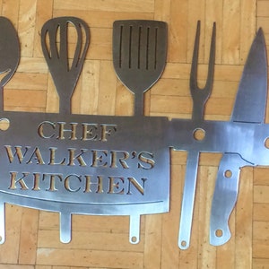 May include: A metal kitchen sign featuring cut-out silhouettes of cooking utensils, including a spoon, whisk, spatula, fork, and knife. The sign reads "CHEF WALKER'S KITCHEN" in a bold, readable font. The sign is made of a silver-colored metal.