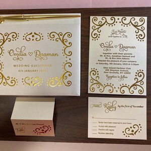 May include: A white wedding guest book with gold foil accents and the text "Omelia & Daaman Wedding Guestbook 4th January 2025".