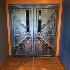 May include: A pair of industrial-style metal doors with a weathered appearance. The doors feature yellow and black diagonal stripes, the word "CAUTION", and other warning symbols. The doors are set in a doorway with orange and blue walls and a wooden floor.