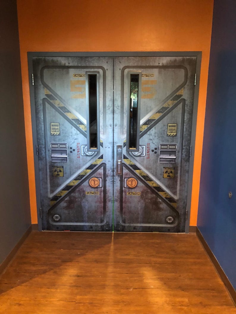 May include: A pair of industrial-style metal doors with a weathered appearance. The doors feature yellow and black diagonal stripes, the word "CAUTION", and other warning symbols. The doors are set in a doorway with orange and blue walls and a wooden floor.