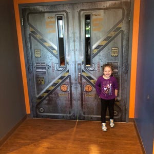 May include: A young girl stands in front of a pair of large, industrial-style doors. The doors are gray with yellow and black hazard stripes and the word "CAUTION" in orange. The girl wears a purple long-sleeved shirt, black pants, and sneakers.