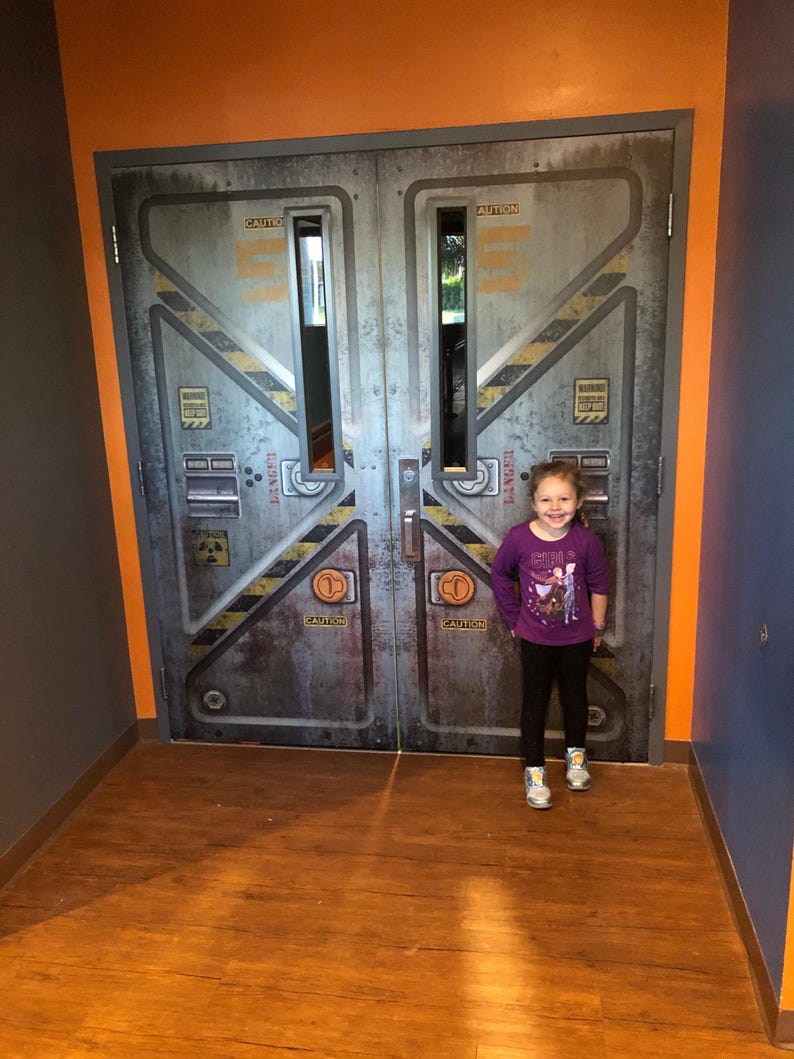 May include: A young girl stands in front of a pair of large, industrial-style doors. The doors are gray with yellow and black hazard stripes and the word "CAUTION" in orange. The girl wears a purple long-sleeved shirt, black pants, and sneakers.