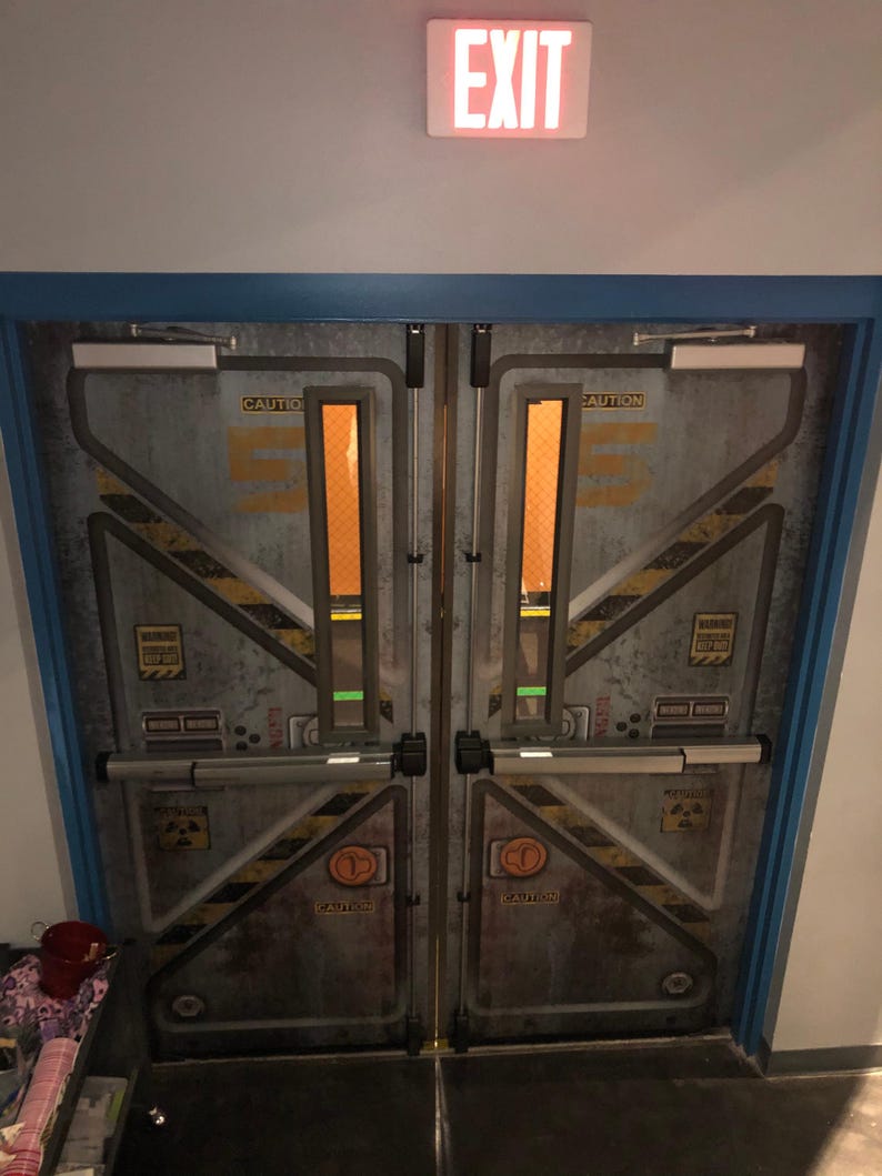 May include: A pair of industrial-style double doors with a weathered metal finish. The doors feature yellow and black hazard stripes, caution labels, and an illuminated "EXIT" sign above. The door frames are trimmed in blue.