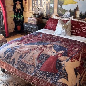 Può includere: Un letto con un copripiumino caratterizzato da una stampa in stile arazzo rosso scuro e blu che raffigura un unicorno e un leone. Il letto include due cuscini rossi con testo bianco e un cuscino decorativo chiaro, vicino a un comodino nero e una mensola.