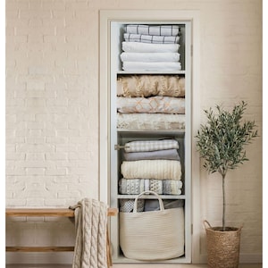 May include: A white closet filled with folded bedding, including sheets, blankets, and decorative pillows. A large woven basket sits at the bottom, and a small olive tree in a woven pot is to the right. A wooden bench and a cable knit blanket are in the foreground.