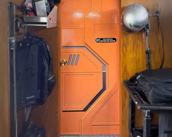 Pode incluir: Uma porta laranja com um design futurista, com detalhes em preto e um pequeno sinal retangular que diz "02 Restricted Access". A porta tem uma maçaneta dourada e uma série de painéis retangulares e angulares. Um capacete está montado na parede.