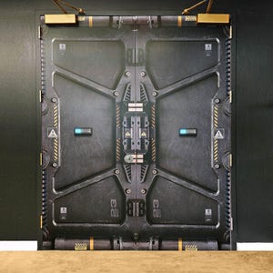 May include: A large, industrial-style door with a metallic, textured surface. The door is primarily dark grey with yellow and blue accents, featuring intricate geometric designs and the number "23". It has a futuristic, sci-fi aesthetic.