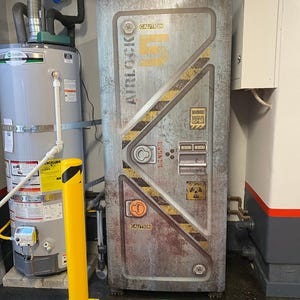 May include: A weathered metal door with the word "AIRLOCK" and the number "5" in yellow. The door has caution labels and diagonal yellow and black stripes. The door is in a utility room with a water heater and a yellow bollard.