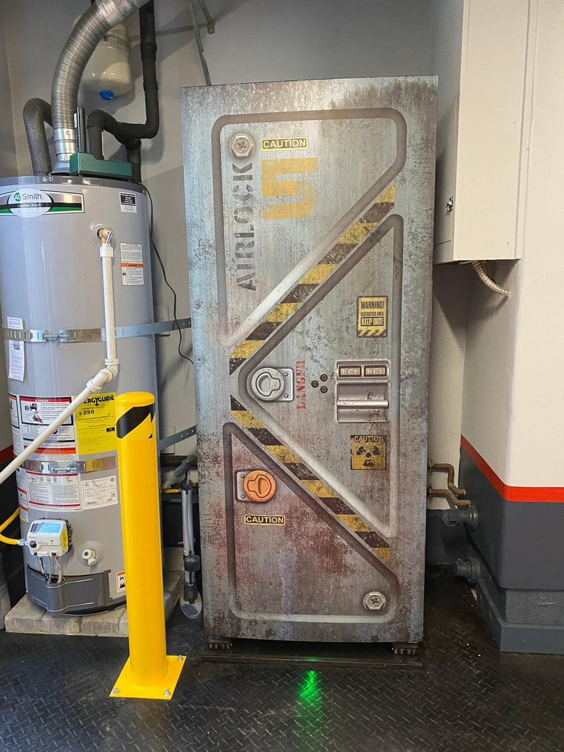 May include: A weathered metal door with the word "AIRLOCK" and the number "5" in yellow. The door has caution labels and diagonal yellow and black stripes. The door is in a utility room with a water heater and a yellow bollard.