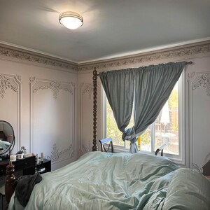 May include: A bedroom with a large bed with a light blue comforter and a light blue headboard. The room has a window with light blue curtains and a white trim. The walls are painted a light pink color and have a decorative molding around the top.