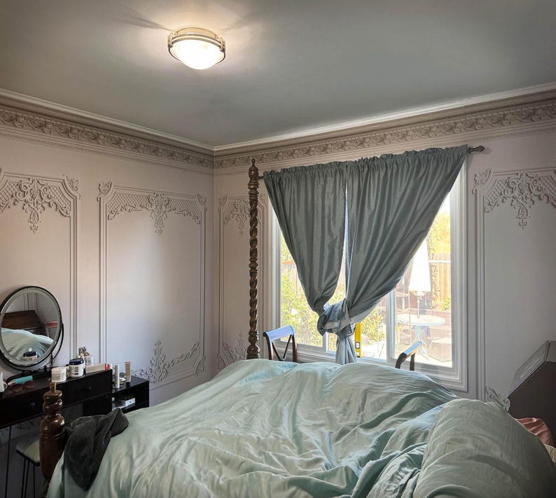 May include: A bedroom with a large bed with a light blue comforter and a light blue headboard. The room has a window with light blue curtains and a white trim. The walls are painted a light pink color and have a decorative molding around the top.