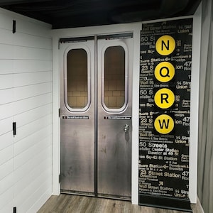 Peut inclure: Une porte de métro décorative avec une finition en métal argenté et une fenêtre rectangulaire. La porte porte une pancarte indiquant "Do not lean on door". Un panneau noir affiche des symboles de lignes de métro jaunes et des noms de stations.