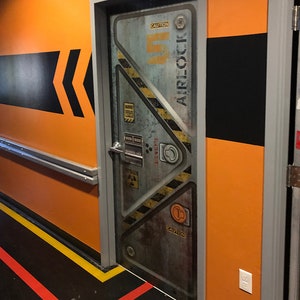 May include: A metallic gray door with the word "AIRLOCK" and the number "5" in a sci-fi style. The door has yellow and black hazard stripes and is set in an orange wall with black and yellow accents. The floor has red and yellow lines.