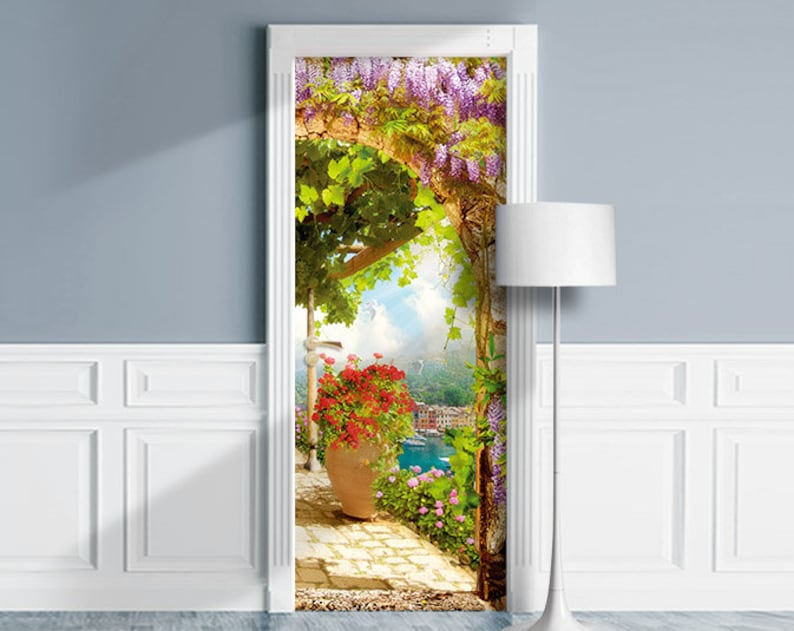 Pode incluir: Um adesivo de porta que apresenta um arco floral colorido com vista para uma cidade costeira. O arco &eacute; coberto de flores de glicinia roxas e videiras verdes. Um vaso vermelho com uma planta fica na frente do arco.