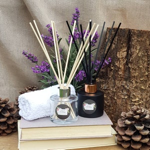 May include: Two glass diffuser bottles with black and natural reed sticks. The bottles have black labels with white text that reads "Enchanted Home Fragrances". One bottle is clear glass with a silver lid and the other is black glass with a copper lid. The bottles are sitting on a stack of books with a white towel and pine cones on either side.