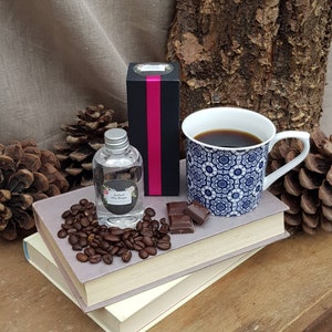 May include: A clear glass bottle with a silver lid and a black and white label that says "Infused Skin Soother". The bottle is sitting on a stack of books with coffee beans and a piece of chocolate. A blue and white mug with coffee is in the background.