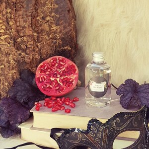May include: A clear glass bottle with a silver cap and a black label that reads "Enchanted Eden Designs". The bottle is sitting on a stack of books with a black and gold masquerade mask in the foreground. There are pomegranate seeds scattered around the bottle.