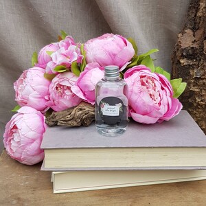 May include: A clear glass bottle with a silver lid filled with a liquid. The bottle has a black label with the text "Ireland Eden Droops" and "Luxury Home Fragrance". The bottle is surrounded by pink peonies and a piece of driftwood. The bottle is sitting on top of a stack of books.