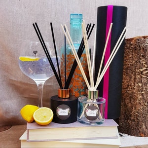 May include: Two black and clear glass diffuser bottles with black and white reeds and a gold lid. The bottles are sitting on a stack of books with a glass of gin and tonic and lemons in the foreground.