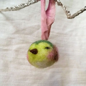 May include: A small, hand-felted bird ornament with a green, yellow, and pink body. The bird is hanging from a pink ribbon.