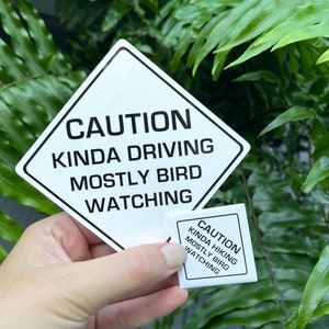 May include: Two white diamond-shaped signs with black text. The larger sign reads "CAUTION KINDA DRIVING MOSTLY BIRD WATCHING". The smaller sign reads "CAUTION KINDA HIKING MOSTLY BIRD WATCHING".