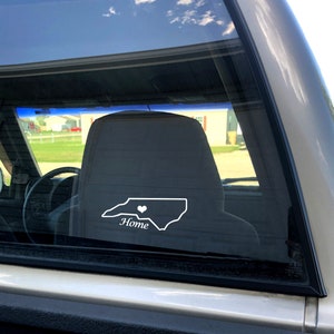 May include: A white vinyl decal of the state of North Carolina with a small heart in the center and the word "Home" underneath. The decal is on the back window of a silver pickup truck.