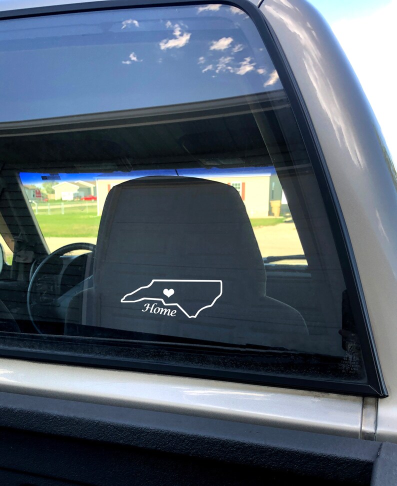 May include: A white vinyl decal of the state of North Carolina with a small heart in the center and the word "Home" underneath. The decal is on the back window of a silver pickup truck.