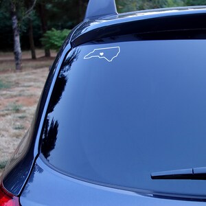 May include: A white vinyl decal of the outline of North Carolina with a small heart in the center. The decal is on the back window of a dark blue car.