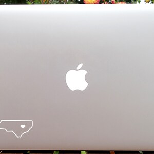 May include: A silver laptop computer with a white Apple logo and a white outline of the state of North Carolina with a heart in the center.