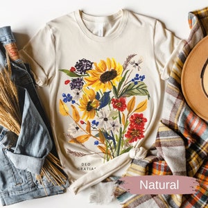 A women's natural colored t-shirt featuring a loose watercolor floral bouquet with yellow sunflowers and berries. The words "Deo Gratias" are at the bottom of the t-shirt. Shirt is styled with a jean jacket, a brown wool hat and plaid scarf For Fall