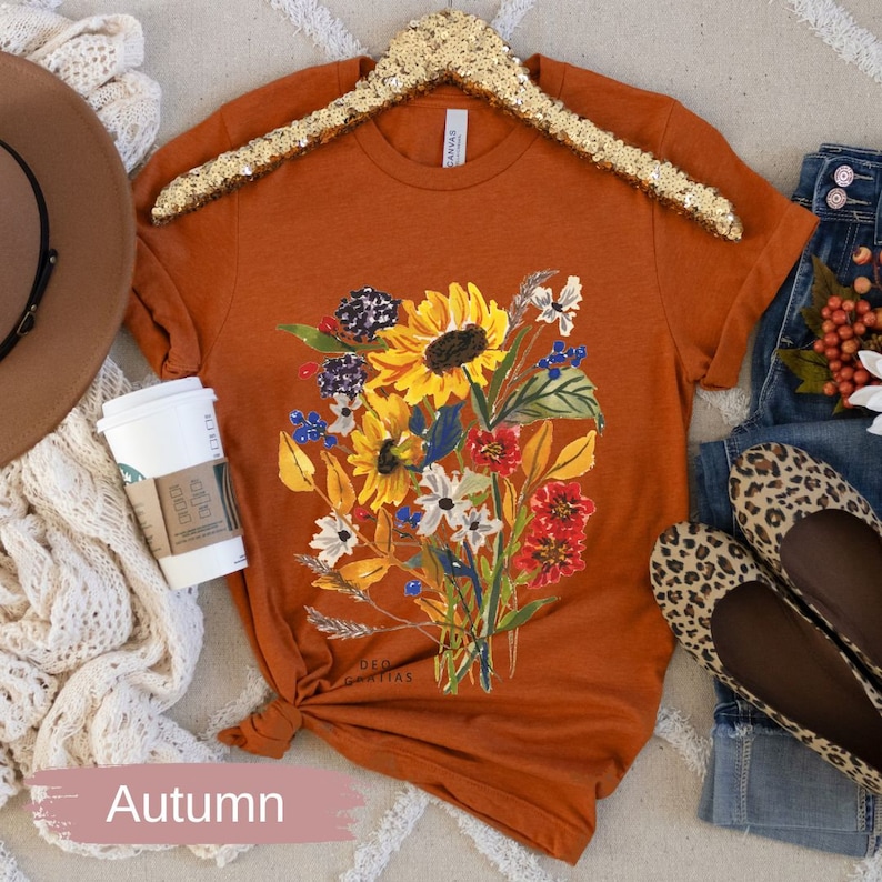 Puede incluir: Una camiseta de color &oacute;xido con un estampado floral que incluye girasoles, flores azules y blancas, y flores rojas y naranjas. El texto "DEO GRATIAS" est&aacute; impreso en la camiseta.
