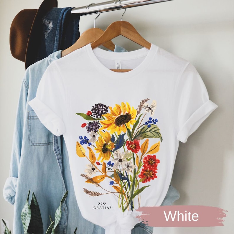Puede incluir: Camiseta blanca con estampado floral de colores con girasoles, flores rojas, amarillas y azules, y hojas verdes. El texto "DEO GRATIAS" est&aacute; impreso en la parte inferior de la camiseta.