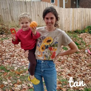 Puede incluir: Una mujer con una camiseta de color canela con estampado floral y pantalones vaqueros azules est&aacute; sosteniendo a un ni&ntilde;o peque&ntilde;o. El ni&ntilde;o lleva un su&eacute;ter rojo y pantalones marrones. La mujer sonr&iacute;e y el ni&ntilde;o sostiene una peque&ntilde;a calabaza naranja.
