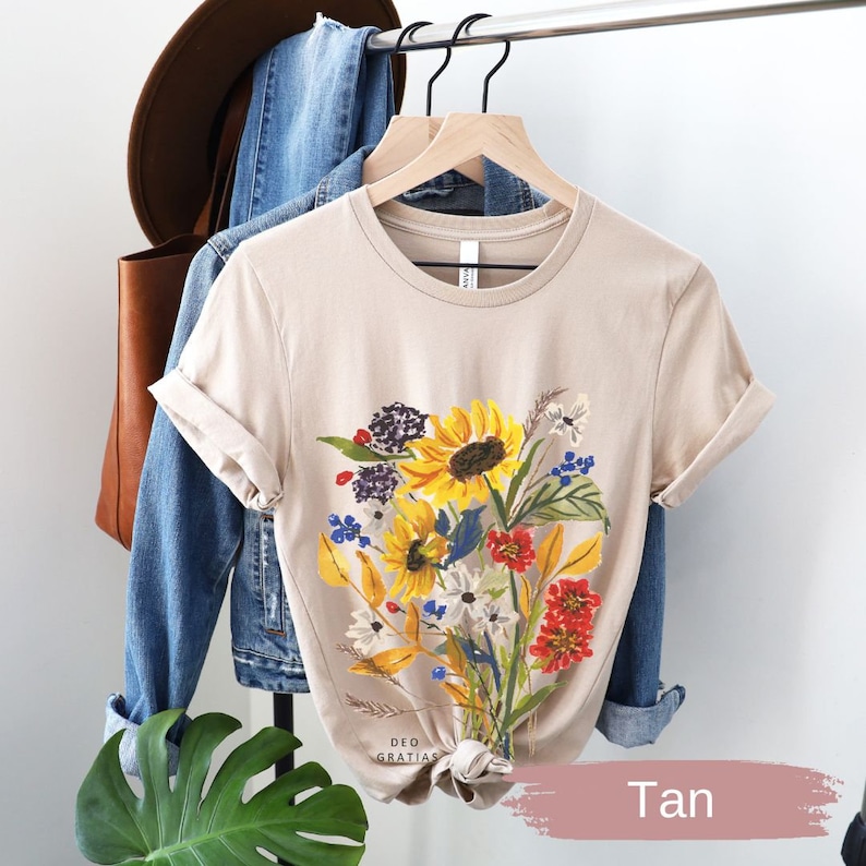 Puede incluir: Camiseta de manga corta de color marr&oacute;n claro con estampado floral de girasoles, flores rojas y blancas y hojas verdes. El texto "DEO GRATIAS" est&aacute; impreso en la parte inferior de la camiseta.