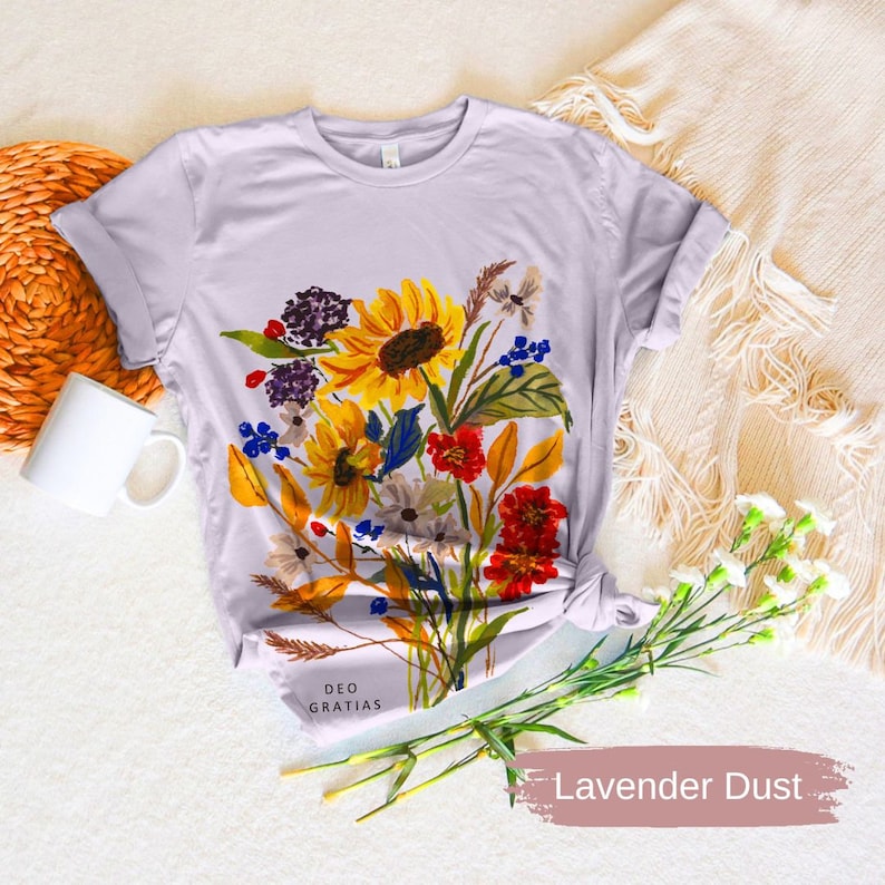 Puede incluir: Una camiseta de color morado claro con un dise&ntilde;o floral que incluye girasoles, margaritas y otras flores silvestres en tonos amarillos, rojos, naranjas y morados. El texto "DEO GRATIAS" est&aacute; impreso en la parte inferior de la camiseta.